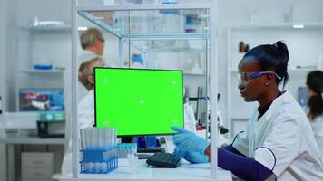 Black woman researcher looking at chroma key computer in modern equipped lab. Multiethnic team of microbiologists doing vaccine research writing on device with green screen, isolated, mockup display. video