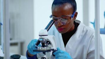Close up of black woman looking in microscope in modern equipped lab. Multiethnic team examining virus evolution using high tech for scientific research of treatment development against covid19 video