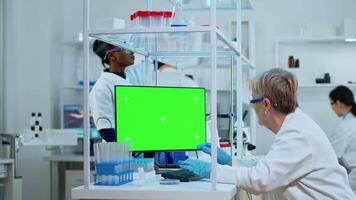 Senior doctor working at computer with green screen in modern equipped lab. Multiethnic team of microbiologists doing vaccine research writing on device with chroma key, isolated, mockup display. video