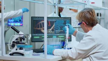 Elderly research scientist using micropipette filling test tubes in modern equipped lab. Multiethnic team examining virus evolution using high tech analysing treatment development against covid19. video