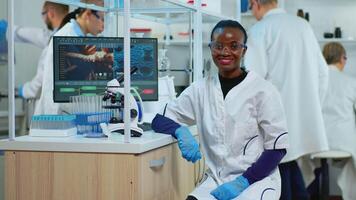 Professional black woman scientist looking at camera smiling in modern equipped lab. Multiethnic team examining virus evolution using high tech and tools for scientific research, vaccine development. video