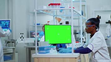 African scientist woman typing on computer with green mockup in modern equipped lab. Multiethnic team of microbiologists doing vaccine research writing on device with chroma key, isolated display. video