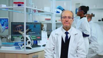 Senior químico sentado dentro moderno equipado laboratório olhando satisfeito às Câmera. multiétnico equipe examinando vírus evolução usando Alto tecnologia e química Ferramentas para científico pesquisar, vacina desenvolvimento video