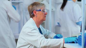 Close up of elderly chemist woman typing on computer in modern equipped laboratory. Multiethnic stuff analysing vaccine evolution using high tech for researching treatment against covid19 virus video
