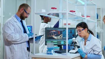 Team of scientists doing biological researches under microscope, lab technician writing data into tablet in modern equipped laboratory. Multiethnic group examining virus evolution using high tech. video