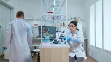 Woman scientist examining petri dish and looking down on microscope in modern equipped laboratory. Multiethnic team analysing vaccine evolution using high tech for researching treatment against virus video