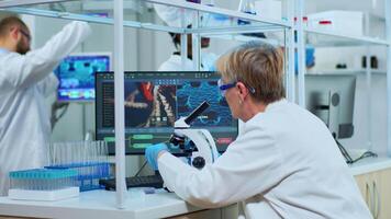 Senior scientist looking through a microscope in modern equipped lab. Multiethnic team examining virus evolution using high tech for scientific research of vaccine development against covid19. video