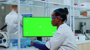 African doctor working at computer with green screen in modern equipped lab. Multiethnic team of microbiologists doing vaccine research writing on device with chroma key, isolated, mockup display. video