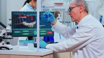 Elderly doctor writing on pc datas from test tubes in modern equipped laboratory. Multiethnic team of doctors examining vaccine evolution using high tech researching diagnosis against covid19 virus video