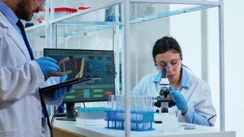 Team medical scientist conducting DNA experiments under digital microscope writing results into tablet working in science laboratory. Caucasian lab engineer in white coat analysing vaccine development video