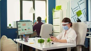 Woman entrepreneur with protective mask working in professional workspace typing on mobile phone in front of computer. Multiethnic coworkers working respecting social distance in financial company video