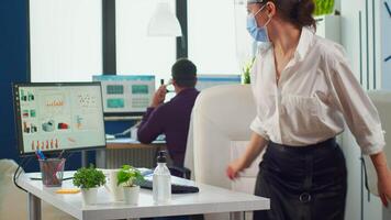 Entrepreneur with protection mask and visor applying sanitizer gel rubbing hands before typing at computer. Businesswoman in new normal workplace disinfecting while colleagues working in background video