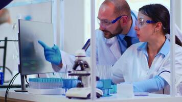 Chemist nurse explainig to doctor vaccine development in modern equipped laboratory pointing on computer desktop. Team of doctors examining virus evolution using high tech researching diagnosis video