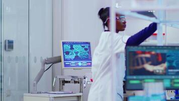 Black young scientist with white coat working in busy modern laboratory. Diverse team of industrial scientists, engineers, developers innovating new vaccine, analysing virus evolution using high tech video