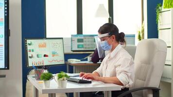 Coworkers with protection face masks working together in workplace during pandemic. Team in new normal business financial office typing on computer, checking reports, analysing data looking at desktop video