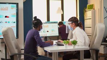 Manager talking with african employee about company statistics wearing protection face mask and visor against coronavirus sitting at desk respecting social distance. New normal business office. video