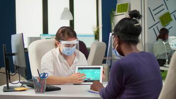 Group of multiethnic confident business people with face masks analyzing data using digital tablet while sitting in office with new normal. Colleagues working in background respecting social distance video