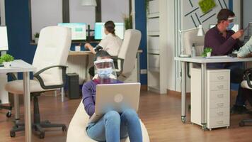 African employee with visor and face mask sitting on armchair in middle of office room working on laptop. Multiethnic business team planning company strategy respecting social distance. video
