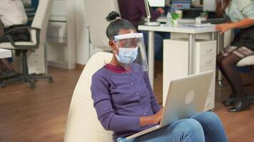Black freelancer with protection mask against coronavirus sitting in middle of workplace office room analysing project on laptop. Multiethnic business team working respecting social distance video