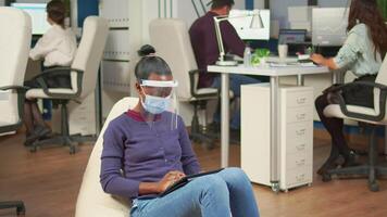 Black freelancer with protection mask against coronavirus sitting on armchair in middle of office room analysing project on digital tablet. Multiethnic business team working respecting social distance video