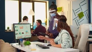 Multiethnic coworkers with protection masks checking annual statistics looking on graphs in computer and clipboard, black woman taking notes on tablet. Diverse team working respecting social distance. video