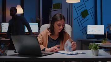 Overworked businesswoman analyzing financial graphs on clipboard comparing with economic statistics using laptop computer. Tired entrepreneur woman working in business company office late at night video