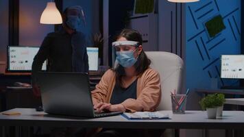 Workaholic tired businesswoman with face mask and visor against covid19 working in company office typing financial statistics on laptop. Manager remains alone in company room after collegue left video