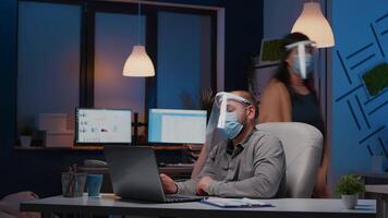 Workaholic businessman with face mask and visor against covid19 working in company office analysing marketing statistics on laptop. Manager remains alone in company room after his collegue left video