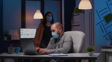 Overworked businessman with face mask against covid19 working in startup office checking management strategy late at night. Exhausted manager remains alone in company room after his collegue left video