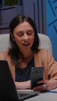 Overworked businesswoman with headphones discussing marketing strategy with teamwork using smartphone. Tired entrepreneur having online videocall financial interview sitting at desk in company office video
