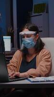Workaholic tired businesswoman with face mask and visor against covid19 working in company office typing financial statistics on laptop. Manager remains alone in company room after collegue left video