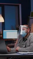 Workaholic businessman with face mask and visor against covid19 working in company office analysing marketing statistics on laptop. Manager remains alone in company room after his collegue left video