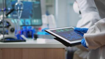 Back view of scientist in coverall analyzing virus evolution looking on digital tablet. Team of microbiologists conducting vaccine development using high tech for researching treatment against covid19 video