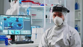 Tired doctor in protection suit against covid-19 looking exhausted at camera in equipped lab. Team of scientists examining virus evolution using high tech for scientific research, vaccine development. video