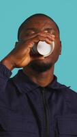 Vertical Headshot portrait of security agent drinking coffee to stop yawning while securing assigned post, studio background. Close up shot of guardian consuming caffeinated beverage, camera B video