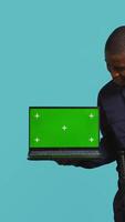 Vertical Portrait of happy security guard at work showcasing green screen laptop while overseeing premises. Cheerful private law enforcement worker presenting chroma key notebook, studio backdrop, camera B video