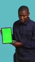 Vertical Portrait of smiling security guard at work showcasing isolated screen tablet while overseeing premises. Joyful private law enforcement worker presenting chroma key device, studio backdrop, camera B video