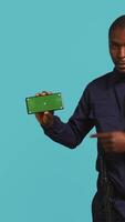 Vertical Joyful security guard working protection job presenting chroma key cellphone. Portrait of merry bodyguard showcasing isolated screen mobile phone, isolated over studio background, camera B video