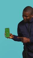 Vertical Portrait of upbeat security staff member showing green screen smartphone. Cheerful patrolling agent maintains secure environment, showcasing mockup phone, studio background, camera B video