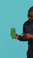 Vertical Portrait of joyous security guard at work showcasing green screen phone while overseeing premises. Jolly private law enforcement worker presenting mockup smartphone, studio backdrop, camera B video
