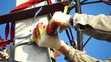 Skilled electrician in helmet fixes wires standing on ladder near high pole against blue sky on summer day backside view. Electrical service and mounting on the pole. Slow motion video