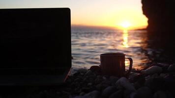 Silhouette of laptop and cup on the beach over sea sunset. Sparkling sea water on background. Freelancer, blogger working on notebook on the beach. video