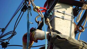Skilled electrician in helmet fixes wires standing on ladder near high pole against blue sky on summer day backside view. Electrical service and mounting on the pole. Slow motion video
