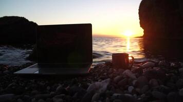Silhouette of laptop and cup on the beach over sea sunset. Sparkling sea water on background. Freelancer, blogger working on notebook on the beach. video