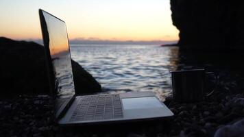 Silhouette of laptop and cup on the beach over sea sunset. Sparkling sea water on background. Freelancer, blogger working on notebook on the beach. video