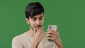 Portrait close-up arabian young man in studio on green background holding phone happy smiling guy browsing social networks make online order read news using modern gadget male customer shopping remote photo