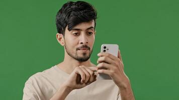 Portrait close-up arabian young man in studio on green background holding phone happy smiling guy browsing social networks make online order read news using modern gadget male customer shopping remote photo
