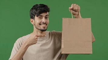 Portrait young smiling guy holding paper package pointing finger to shopping bag hispanic male buyer standing in studio on green background pleased happy man customer enjoying with successful purchase photo