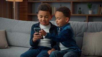 Two little African American siblings boys children kids little brothers with mobile phone smartphone playing game online talking browsing sit at couch at home with cellphone app scrolling web page photo