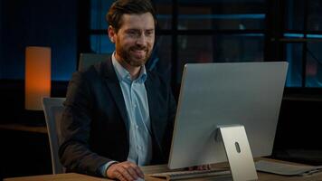 Middle-aged 40s businessman sit in night office company evening online job on computer business project startup. Caucasian adult man employer entrepreneur manager smiling satisfied smile good result photo